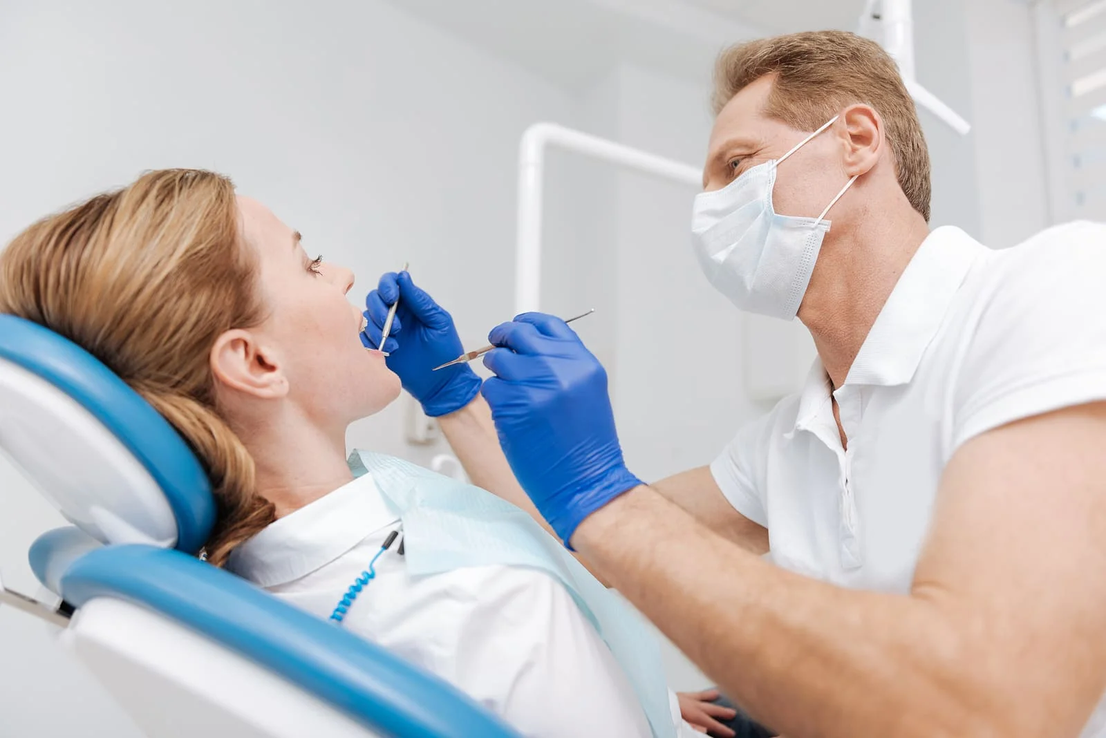 Male dentist in a mask and blue gloves examining a female patient's teeth.